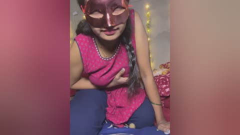 Media: Video of a South Asian woman in a bright pink blouse and blue pants, wearing a red Venetian mask, sitting indoors with fairy lights in the background.