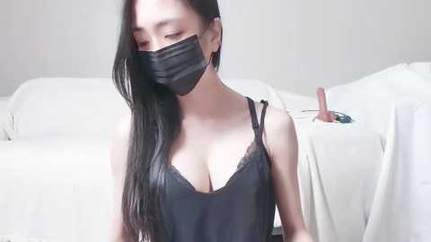 Media: Video of an Asian woman with long black hair, wearing a black face mask and revealing black lace lingerie, sitting on a white couch in a minimalist room.