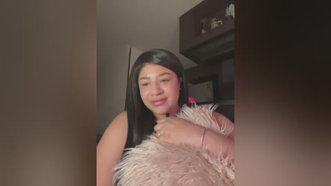 Media: A video of a Latina woman with long black hair, fair skin, and a soft smile, wearing a pink feather boa. She stands in a dimly lit room with shelves in the background.