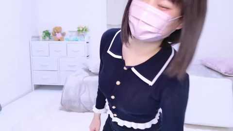 Media: Video of an East Asian woman with light skin and straight brown hair, wearing a navy blue maid outfit with white trim, a pink face mask, and a white apron, standing in a white room with a white dresser, stuffed toys, and a bed in the background.