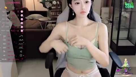 Media: A video of an East Asian woman with pale skin and long black hair, wearing a green camisole and white panties, sitting in a gaming chair in a modern living room.