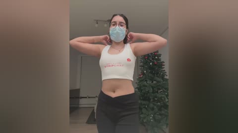 Media: Video of a young woman with a medium build, wearing a white crop top and black pants, adjusting her mask. Background features a Christmas tree and a white wall.