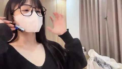 Media: Video of an Asian woman with long black hair, wearing glasses and a white face mask, applying blue and white toothpaste to her teeth in a bathroom with beige curtains and toiletries.