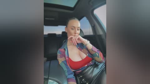 Video of a blonde woman with fair skin and medium breasts, wearing a colorful plaid shirt, red bra, and black leather pants, sitting in a car.