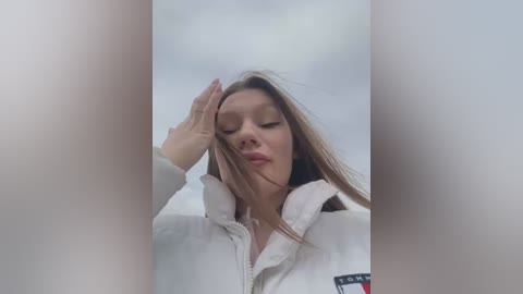 Media: A video captures a young woman with fair skin and long, straight brown hair, wearing a white zip-up jacket. She shields her eyes from the sun with her left hand, her expression neutral. The background is a cloudy sky.