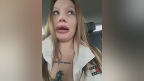 Media: Video of a blonde woman with pale skin, wearing a white jacket, with tattoos on her chest and arm, looking surprised.