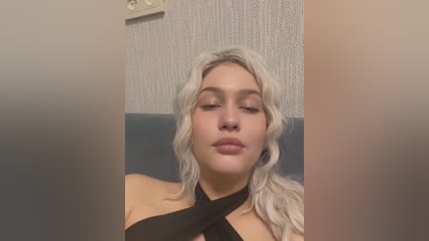 Media: A video of a light-skinned woman with wavy blonde hair, wearing a black halter top, resting her eyes closed in a relaxed pose against a textured gray wall.