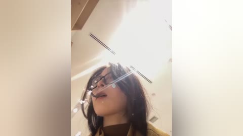 Media: A video of a woman with long, dark hair and glasses, wearing a yellow sweater, smiling slightly, taken from a low angle, indoors with bright lighting.