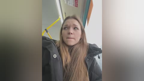 Media: A video shows a Caucasian woman with long, straight, light brown hair, wearing a black winter coat, sitting in a public bus. She looks serious and focused, with a neutral expression.