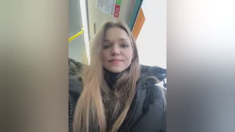 A video of a young woman with long, straight, light brown hair, wearing a black jacket, standing in a brightly lit subway car. The background features colorful posters and a yellow handrail.