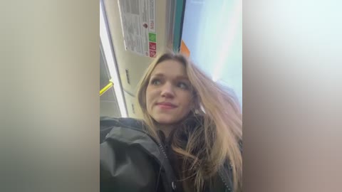 Video of a young woman with long blonde hair in a subway car, wearing a black jacket, smiling. Background features a light-colored wall and a bright, glowing sign.