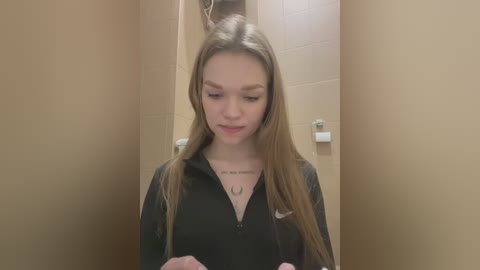 Media: Video of a young Caucasian woman with long, straight brown hair, wearing a black hoodie, standing in a beige-tiled restroom with a toilet roll holder in the background.