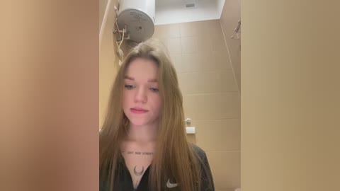 Video of a young Caucasian woman with long, straight blonde hair, wearing a black Nike sweatshirt, standing in a beige-tiled bathroom.