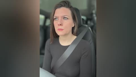 A video of a woman in a car, with shoulder-length brown hair, wearing a black long-sleeve top, and a seatbelt across her chest. The background is blurred, showing a gray interior.