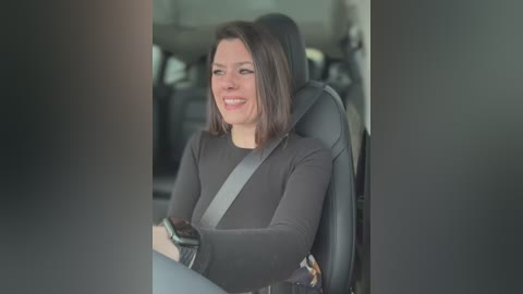 Video of a woman with straight, shoulder-length brown hair, smiling, wearing a black long-sleeve top, seated in a car with a gray seatbelt, blurred background.