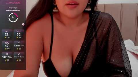 Media: Video of a woman with medium skin tone and long dark hair, wearing a black, semi-sheer blouse with a deep V-neck, revealing ample cleavage. Background features a white bed and beige headboard.