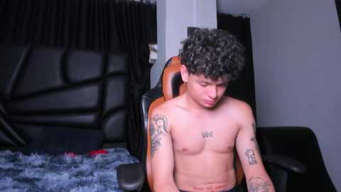 Media: Video of a shirtless, curly-haired young man with tattoos on his arms, sitting in a black gaming chair with a blue blanket on the floor in a dimly lit room.