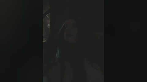 A dimly lit video of a woman with long hair, wearing a dark shirt, standing in a shadowy room with a window partially visible.