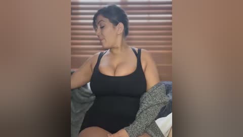 Media: Video of a curvy, Asian woman with short black hair, wearing a black tank top, sitting on a bed with gray and white blankets. Background includes wooden blinds and beige walls.