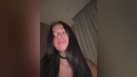 A blurry video of a young woman with long black hair, wearing a black top, standing in a dimly lit room with beige walls and curtains.