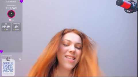 Media: Video of a smiling, fair-skinned woman with long, wavy, reddish-brown hair, captured from a low angle, surrounded by live streaming app interface elements, including a QR code.