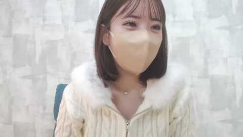 Media: Video of an Asian woman with straight, shoulder-length dark hair, wearing a beige, fur-trimmed jacket, face mask, and light makeup, sitting against a patterned, light-colored wall.