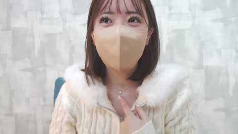 Media: Video of a young East Asian woman with short brown hair, wearing a beige face mask and a cream-colored, fur-trimmed jacket, set against a textured, white and gray abstract backdrop.