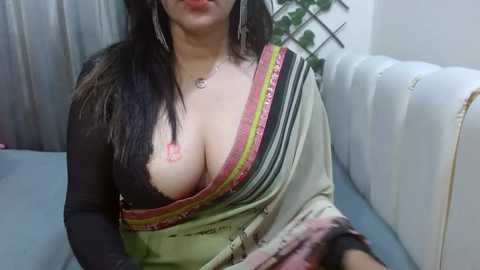 Media: Video of a South Asian woman with fair skin and long black hair, wearing a black see-through blouse revealing her large breasts, seated on a white leather couch, indoors.