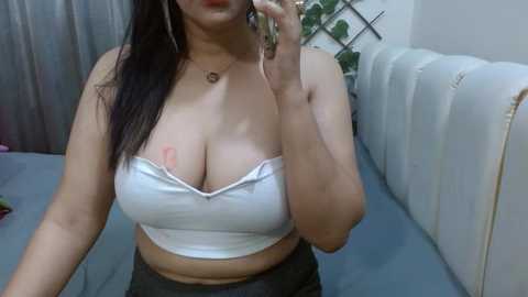 Video of a woman with long dark hair, wearing a white strapless top, revealing ample cleavage, and holding a phone. She stands indoors near a white couch and plant.