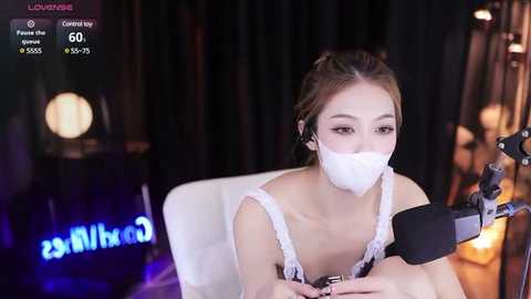 Media: Video of an East Asian woman with light skin and brown hair in a white lace top, wearing a white face mask, recording on a microphone. Background shows dimly lit room with blurred decor.