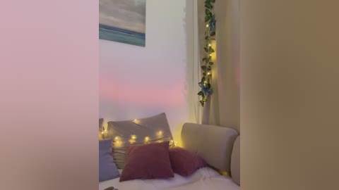 Media: Video of a cozy bedroom with a white bed, gray pillows, pink throw pillows, fairy lights, a green plant, and a seascape painting on the wall.