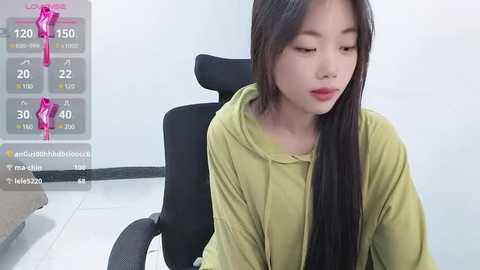 Media: A video of an East Asian woman with long black hair, fair skin, and a slender build, sitting on a black office chair, wearing a light green hoodie, in a minimalist, white-tiled room.