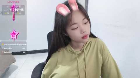 Media: A video of a young Asian woman in a green shirt, wearing pink bunny ears, seated on a black chair, in a brightly lit room with white walls and a gray floor.