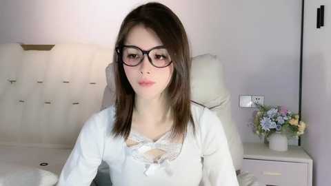 Media: Video of a young Asian woman with straight brown hair, wearing glasses and a white lace-trimmed top, sitting on a white tufted headboard bed with a floral arrangement on a nightstand.