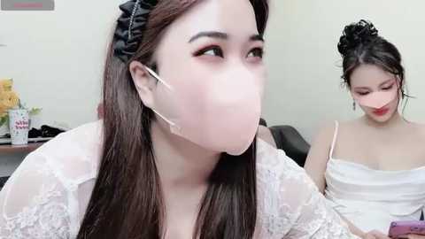 Media: Video of two Asian women with fair skin. One in white lace dress, black headband, pink mask, and headphones, looks thoughtful. The other in white dress, dark hair up, red lipstick, smiles. Background: office with yellow flowers.