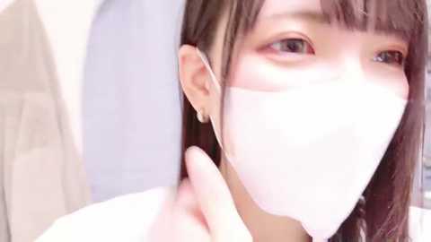 Media: A close-up video of an Asian woman with fair skin, brown hair, and bangs, wearing a white face mask and silver earrings, gazing slightly to the left, in a bright, clean indoor setting.