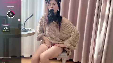 Media: A video of an Asian woman with long black hair, wearing a beige off-shoulder knit sweater, sitting on a white stool, gagged with a black ball gag, and being filmed in a dimly lit room.