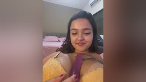 Media: A video of a Latina woman with long black hair, medium skin tone, and large breasts, wearing a yellow lace bra, holding a purple dildo, in a bedroom with a pink bed and air conditioner.