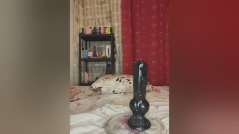 Media: Video of a bedroom with a black dildo on a bedspread, beige and floral-patterned pillows, red curtains with a floral pattern, and a black shelf holding colorful sex toys.