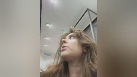 Media: Video of a woman with wavy brown hair, light skin, and a contemplative expression, gazing upwards in an elevator with modern ceiling lights and metal railings.