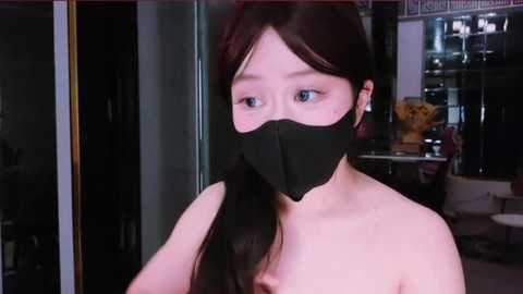 Media: Video of an Asian woman with long black hair, wearing a black mask, topless, in a modern indoor setting with glass walls, reflective surfaces, and a floral arrangement.