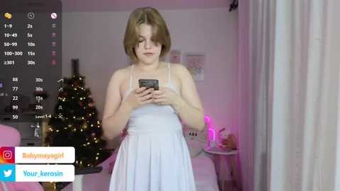 Video of a young woman with short brown hair, wearing a white dress, standing in a brightly lit room with a decorated Christmas tree and pink curtains.