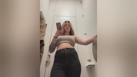 Media: Video of a young woman with fair skin, medium build, and shoulder-length blonde hair, wearing a beige crop top and high-waisted black jeans, taking a selfie in a narrow, white-walled dressing room.