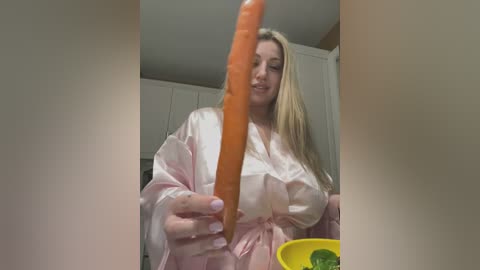 Media: Video of a blonde woman in a pink robe holding a large carrot vertically. She's smiling. Background shows a kitchen with a white cabinet and a yellow bowl containing greens.