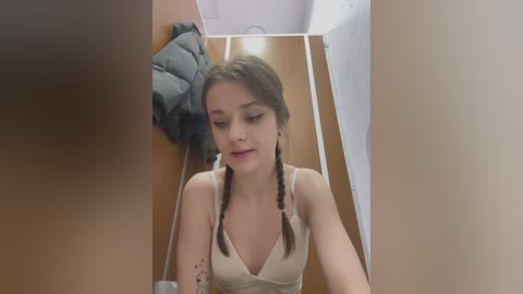 Video of a young Caucasian woman with fair skin, brown hair in twin braids, wearing a beige tank top. She's in a narrow, beige-walled hallway with a gray jacket hanging on a hook.