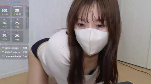 Media: Video of a young Asian woman with straight, shoulder-length brown hair, wearing a white mask, white shirt, and black skirt, leaning forward, with a weather app displaying rain and temperature in the background.