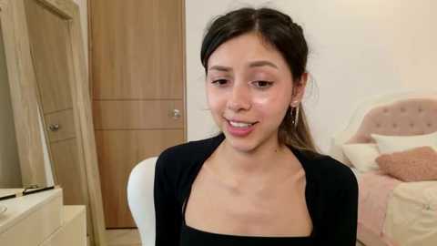 Media: Video of a young woman with light brown skin, long black hair, and small earrings, wearing a black top, seated in a bedroom with wooden furniture and a bed with a beige blanket.