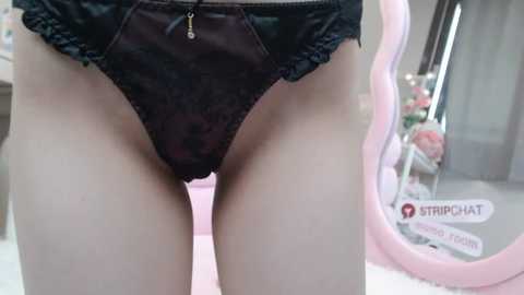 Media: Video of a person wearing black lace panties with a gold charm, standing indoors near a pink-framed mirror and a floral arrangement.