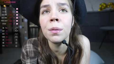 Video of a young woman with light skin, long brown hair, wearing a black hat and plaid shirt, looking worried. Background includes a table with yellow fruit and a chair.