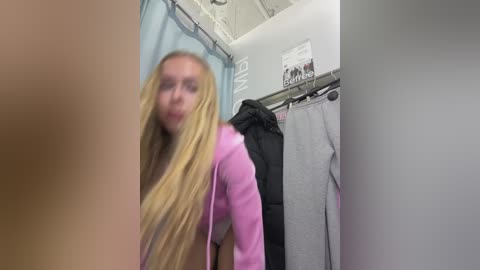 Media: A blurry video of a blonde woman with long hair in a pink jacket, standing in a changing room with gray and black clothes on hangers.